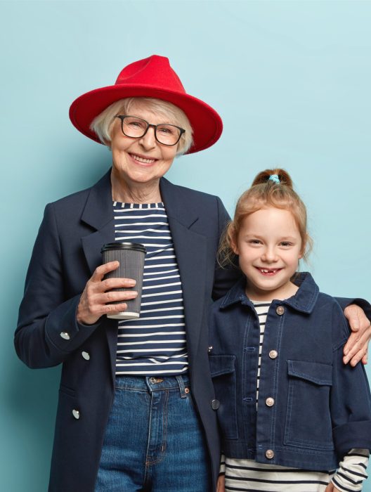 Horizontal view of cheerful grandmother wears red stylish hat, black jacket, drinks takeaway coffee, embraces glad small granddaughter, pose together for family portrait over blue studio wall